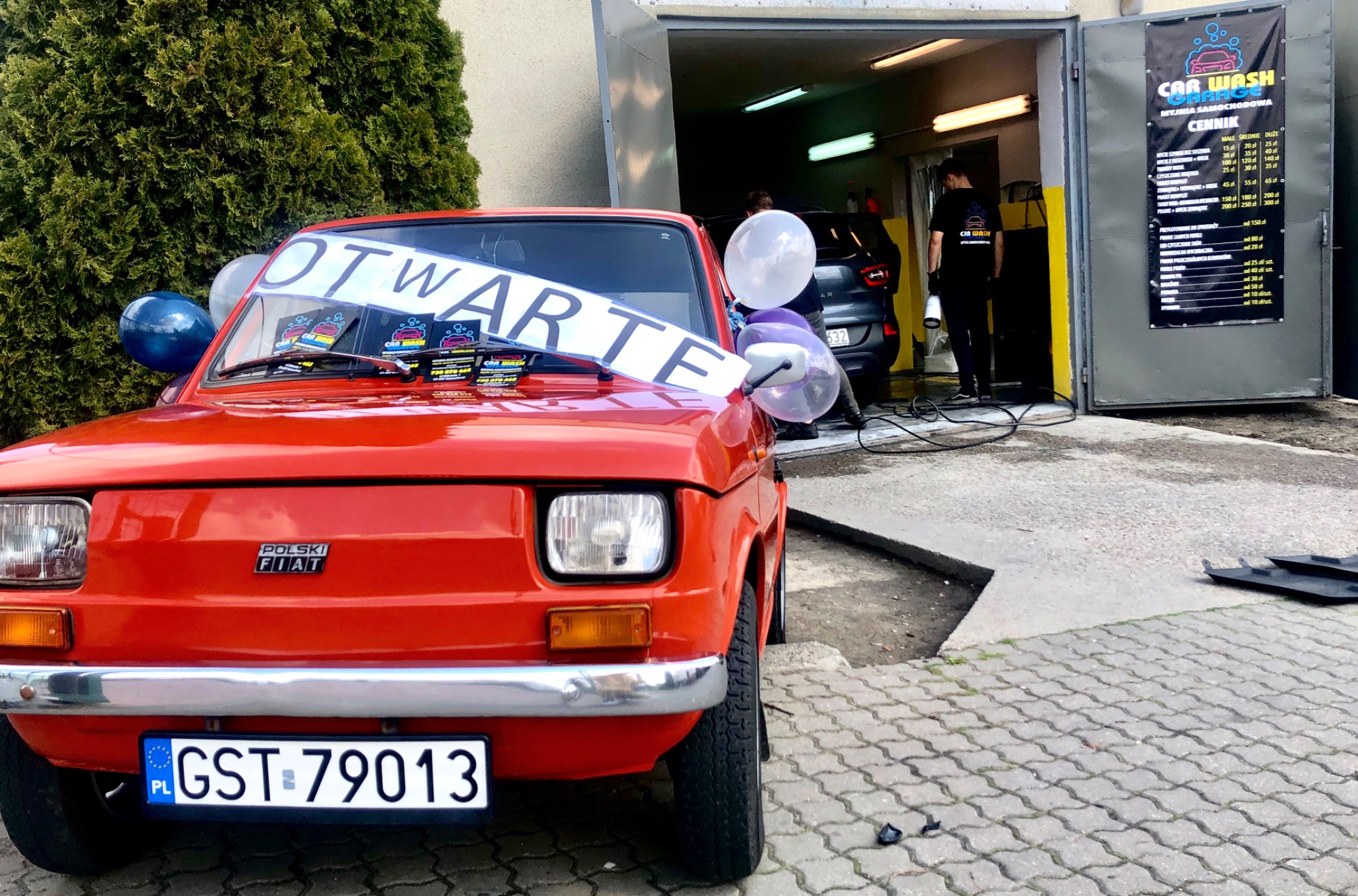 Czerwony Fiat 126p z napisem 'OTWARTE' i balonami przed wjazdem do myjni samochodowej Car Wash Garage, w tle widoczny cennik usług.