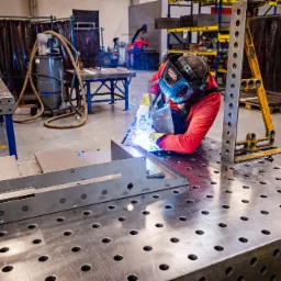 Pracownik w czerwonym kombinezonie spawalniczym i masce ochronnej łączy metalowe elementy na stole spawalniczym z otworami, w tle widoczne stanowiska pracy i regały z metalowymi konstrukcjami.