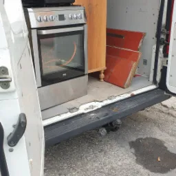 Wnętrze białego busa z otwartymi drzwiami, załadowane sprzętami AGD i meblami: kuchenka, szafka na nóżkach i czerwone drzwi od szafy.