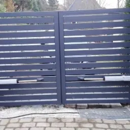 Nowoczesna, pozioma brama wjazdowa z automatycznym napędem, szara, z drobnym żwirem u podstawy i zielenią w tle.