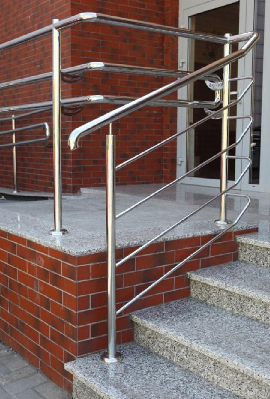 Polerowana, chromowana barierka ze stali nierdzewnej z poziomymi prętami na schodach wejściowych z szarego granitu i ceglaną ścianą w tle.