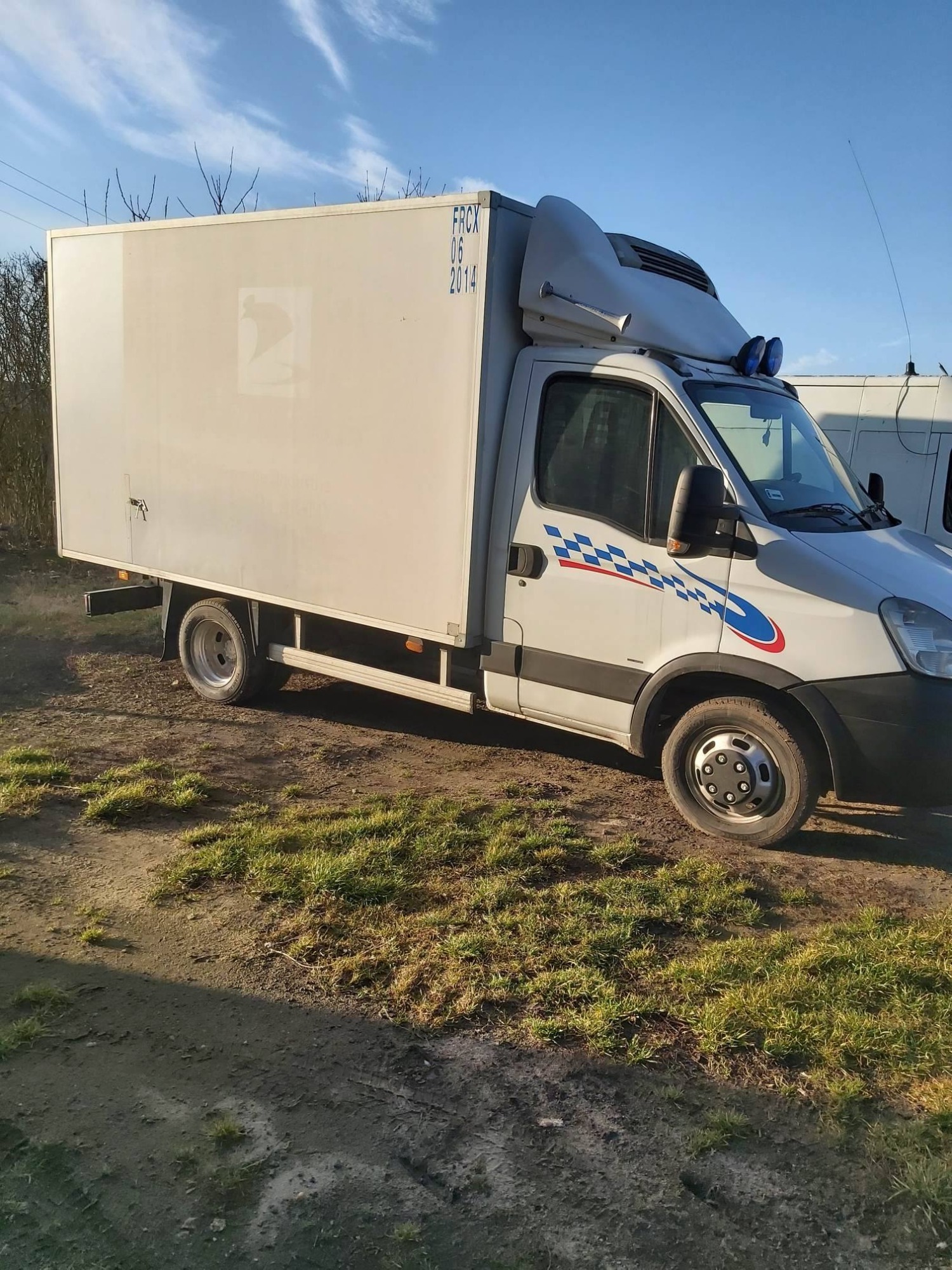 Biały bus chłodnia marki Iveco z niebiesko-czerwonym motywem szachownicy na boku, zaparkowany na trawiastym terenie w słoneczny dzień.