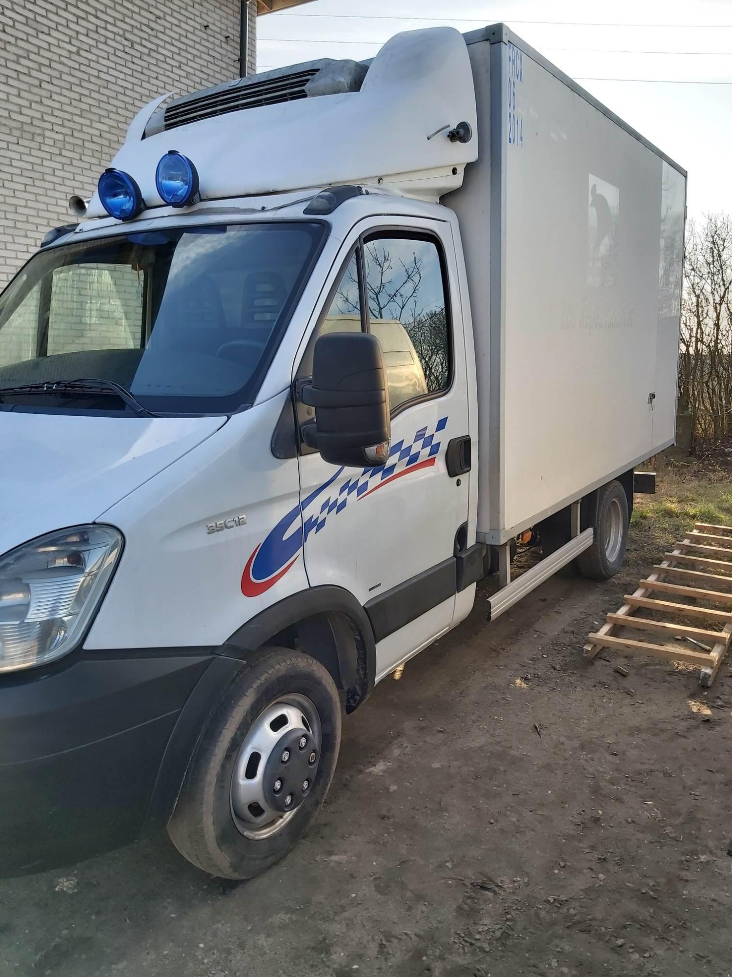 Biały samochód dostawczy z chłodnią, ozdobiony szachownicą, zaparkowany na ziemi obok drabiny z drewna.