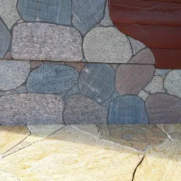 Fragment elewacji z nieregularnych kamieni w odcieniach szarości i różu, połączonych z drewnianą okładziną, oraz podłoga wyłożona płytami z jasnego kamienia z widocznymi spękaniami.