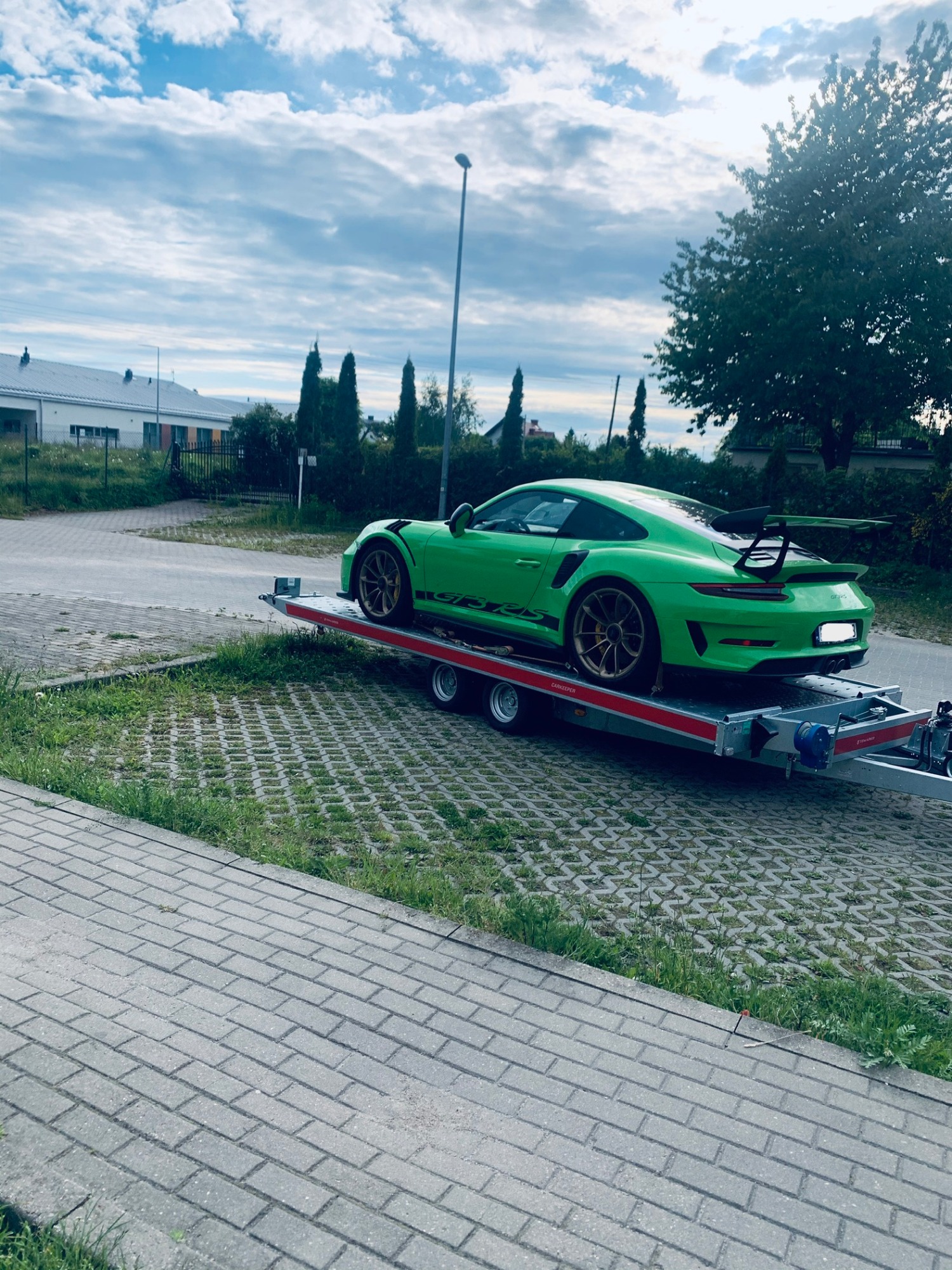 Zielone Porsche GT3 RS na niskopodwoziowej lawecie, widok z tyłu pod kątem, na tle miejskiego krajobrazu z budynkami i drzewami.