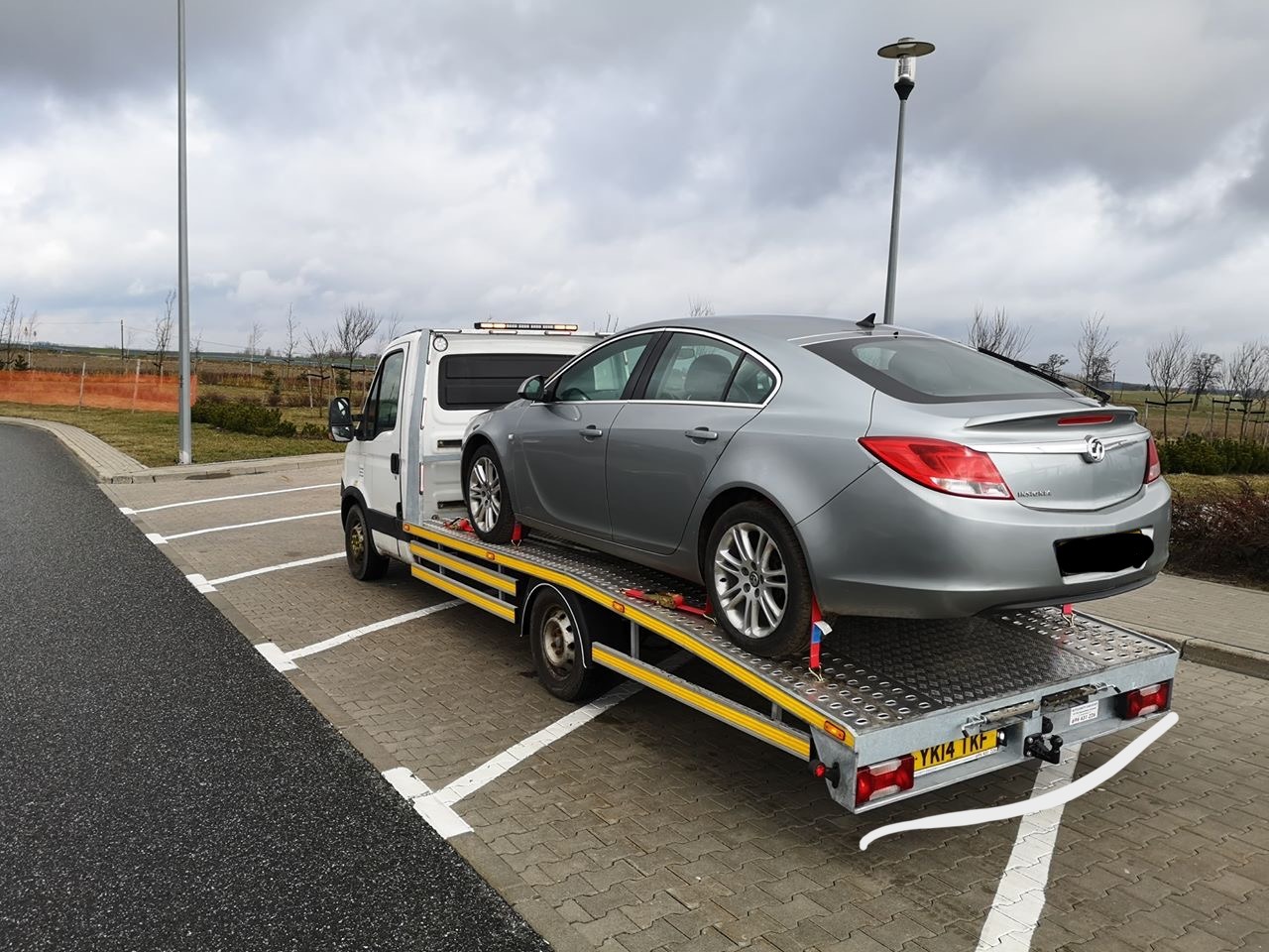 Srebrny samochód osobowy marki Opel Insignia zabezpieczony pasami na żółtej platformie lawety na parkingu z kostki brukowej w pochmurny dzień.