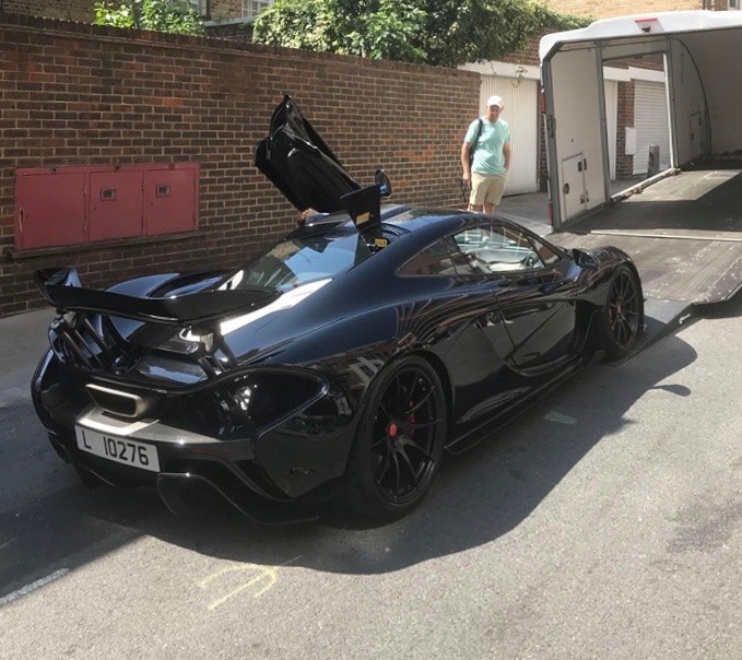 Czarny, sportowy McLaren P1 z otwartymi drzwiami typu 'butterfly' stoi na asfalcie obok lawety z opuszczoną rampą, w tle ceglany budynek i mężczyzna.