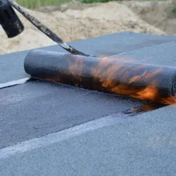 Prace dekarskie: Rozwijanie i zgrzewanie papy termozgrzewalnej na dachu za pomocą palnika dekarskiego, widoczny but roboczy dekarza, w tle piasek.