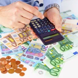 Ręka mężczyzny w błękitnej koszuli i zegarku na bransolecie naciska kalkulator leżący na stosie banknotów euro o różnych nominałach, obok leżą monety