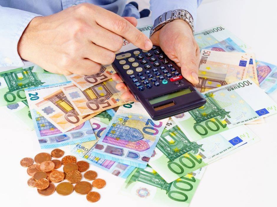 Ręka mężczyzny w błękitnej koszuli i zegarku na bransolecie naciska kalkulator leżący na stosie banknotów euro o różnych nominałach, obok leżą monety