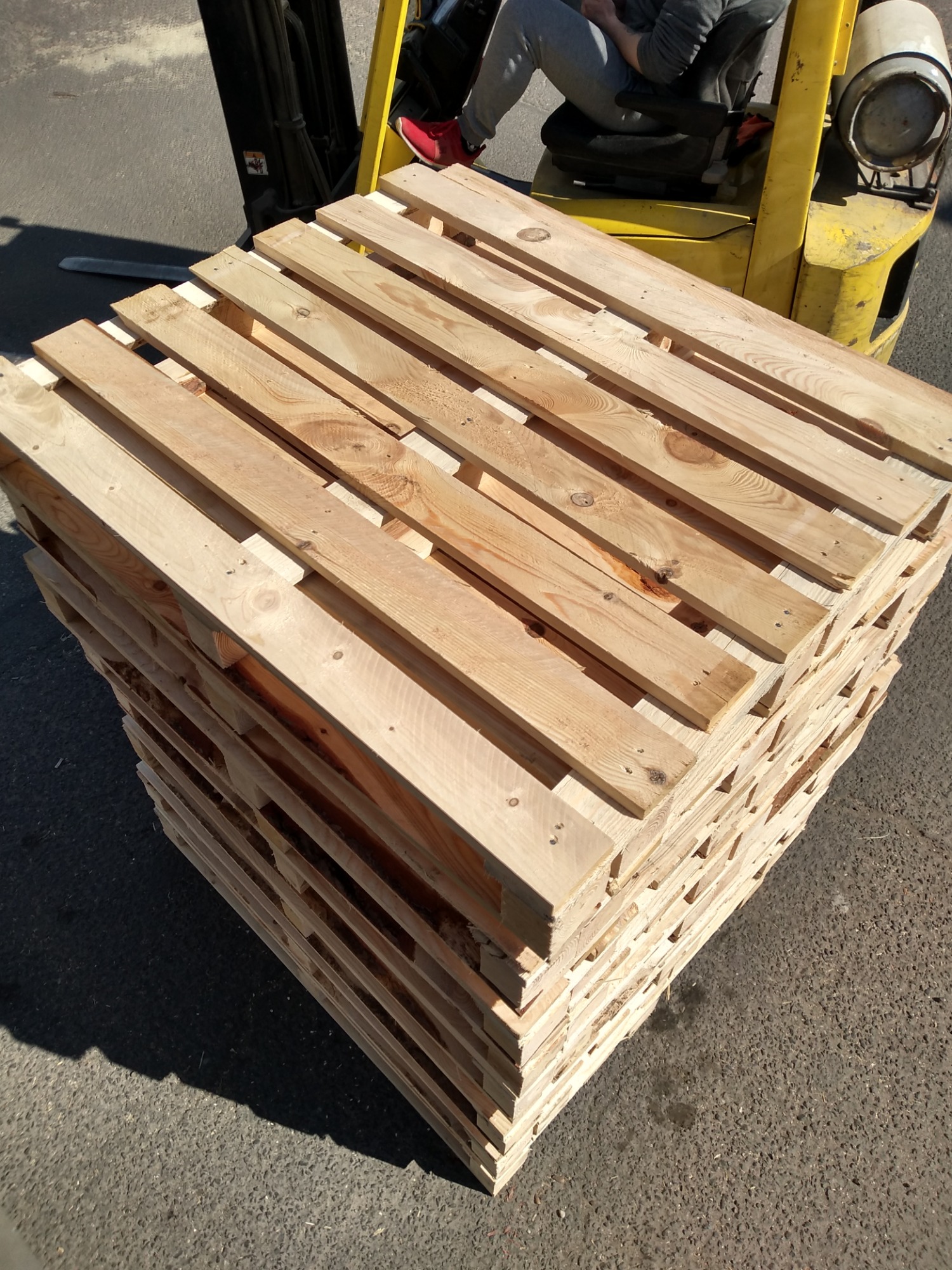 Stos jasnych, drewnianych palet euro ułożonych na zewnątrz, z widocznym w tle fragmentem żółtego wózka widłowego i nogą operatora w czerwonym bucie.