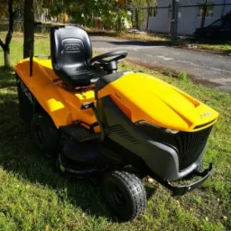 Żółto-czarny traktorek do koszenia trawy marki Stiga na trawniku, widok z przodu, w tle drzewo i budynek.