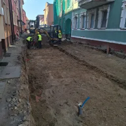 Ulica w trakcie prac ziemnych: mini koparki i robotnicy w kamizelkach odblaskowych przygotowują wykop pod instalacje w zabudowie miejskiej.
