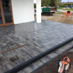 Ułożona, wilgotna kostka brukowa w odcieniach szarości przed budynkiem z widocznymi schodami, obrzeżami, piłą do cięcia kamienia i betoniarką w tle.