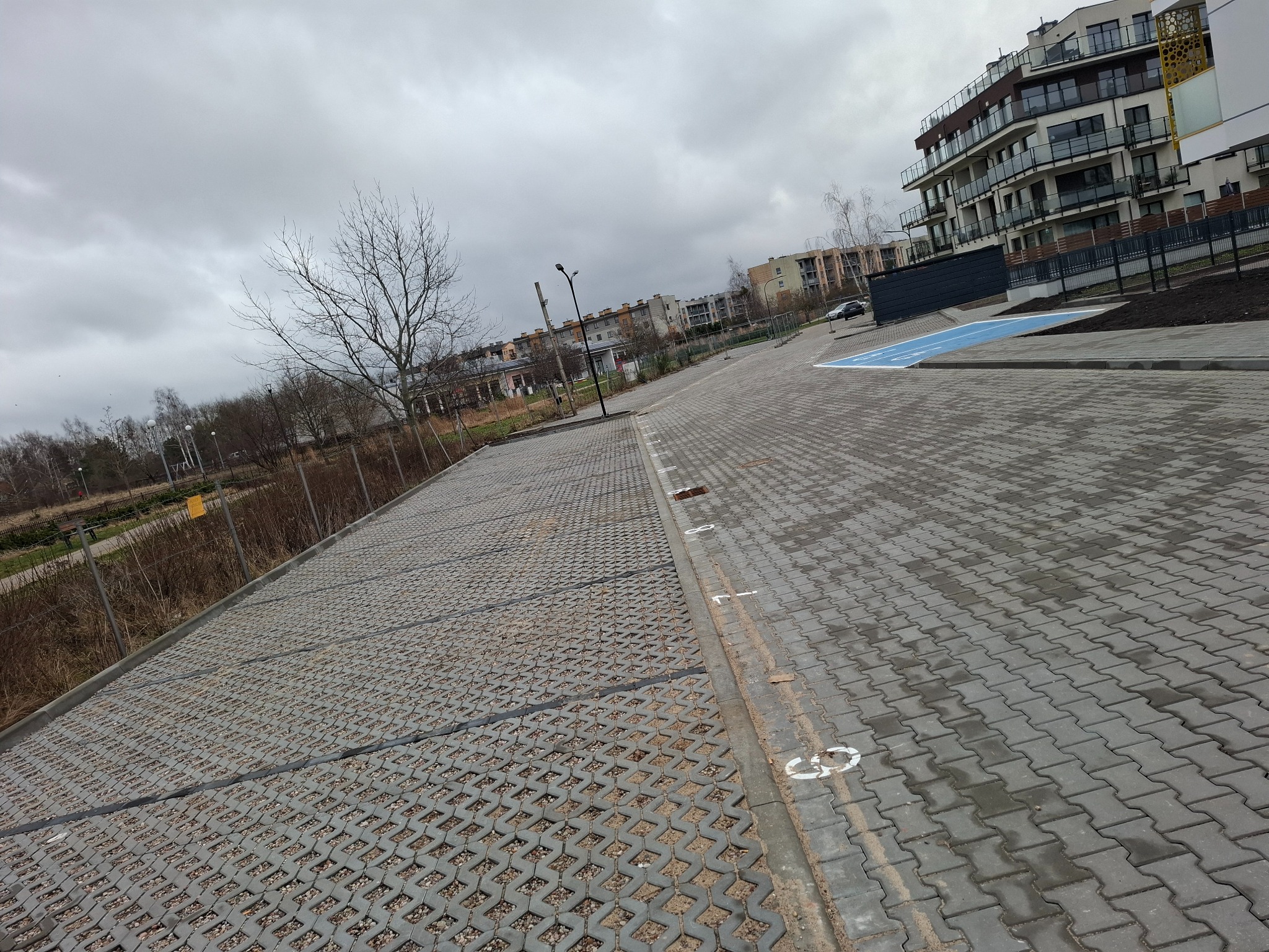 Szary parking z ażurowych płyt betonowych wypełnionych żwirem, z widocznymi numerami miejsc parkingowych namalowanymi na kostce brukowej, w tle budynki mieszkalne pod pochmurnym niebem.