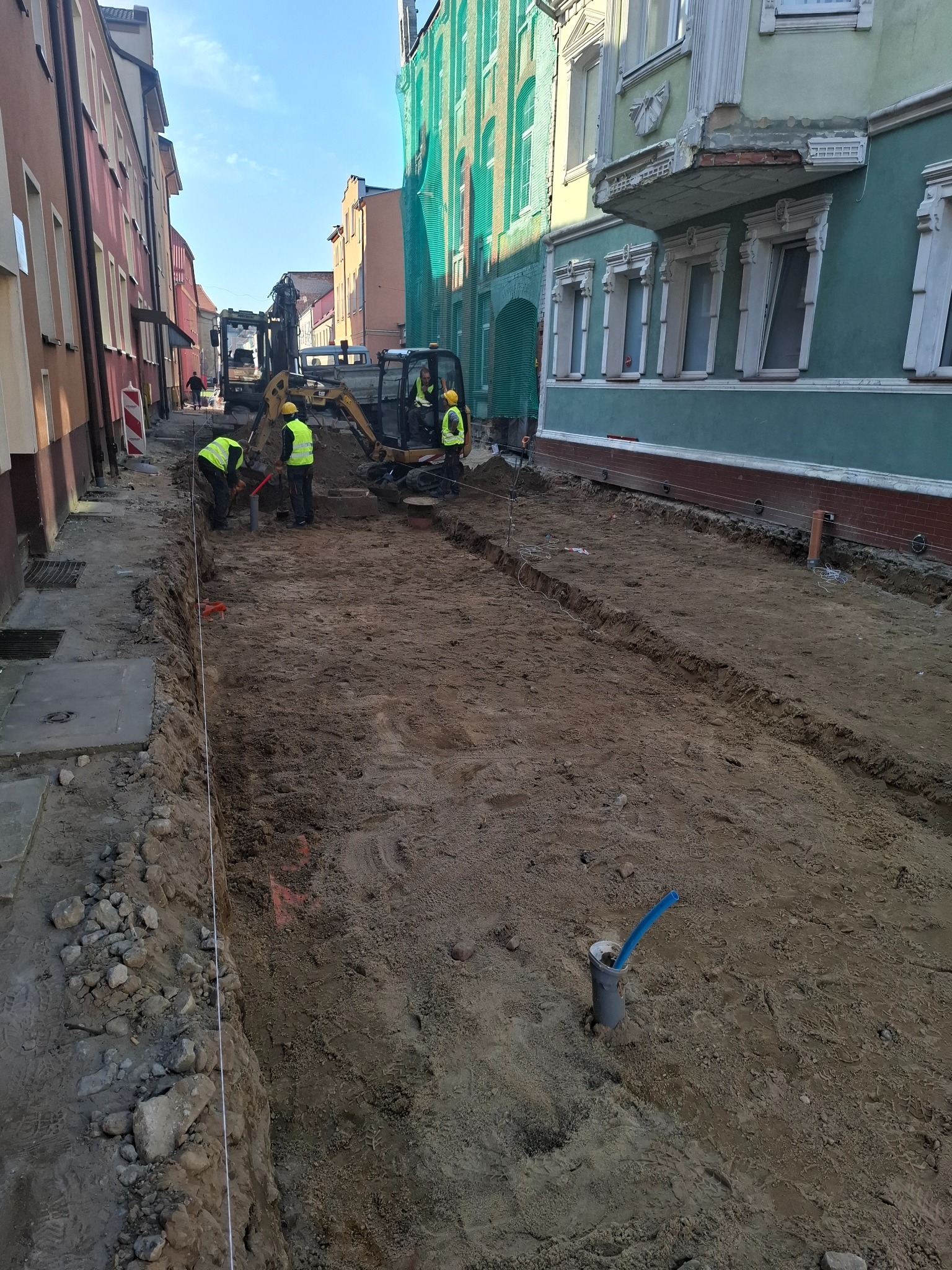 Ulica w trakcie prac ziemnych: mini koparki i robotnicy w kamizelkach odblaskowych przygotowują wykop pod instalacje w zabudowie miejskiej.