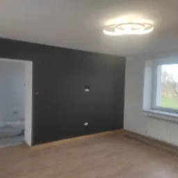 Pomalowane na czarno ściany w pokoju z drewnianą podłogą, widoczny grzejnik pod oknem i nowoczesna lampa sufitowa, w tle wejście do innego pomieszczenia w trakcie prac wykończeniowych, z widocznym...