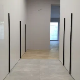 Korytarz z białymi szafami w zabudowie z czarnymi, pionowymi uchwytami, szara podłoga z płytek, parkiet w przejściu do pokoju z oknem z luksferami.