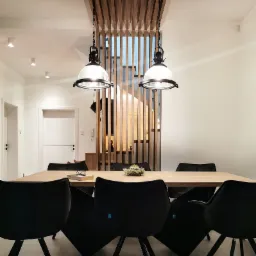 Jadalnia z drewnianym stołem, czarnymi krzesłami i lampami w stylu industrialnym, drewniane lamele na suficie i ścianie, książka i okulary na stole.