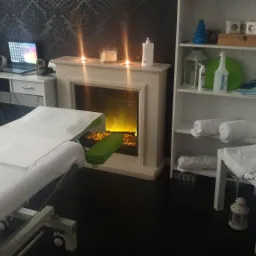 Jasne wnętrze gabinetu z łóżkiem do zabiegów, kominkiem elektrycznym z płomieniami i biurkiem z laptopem w tle.