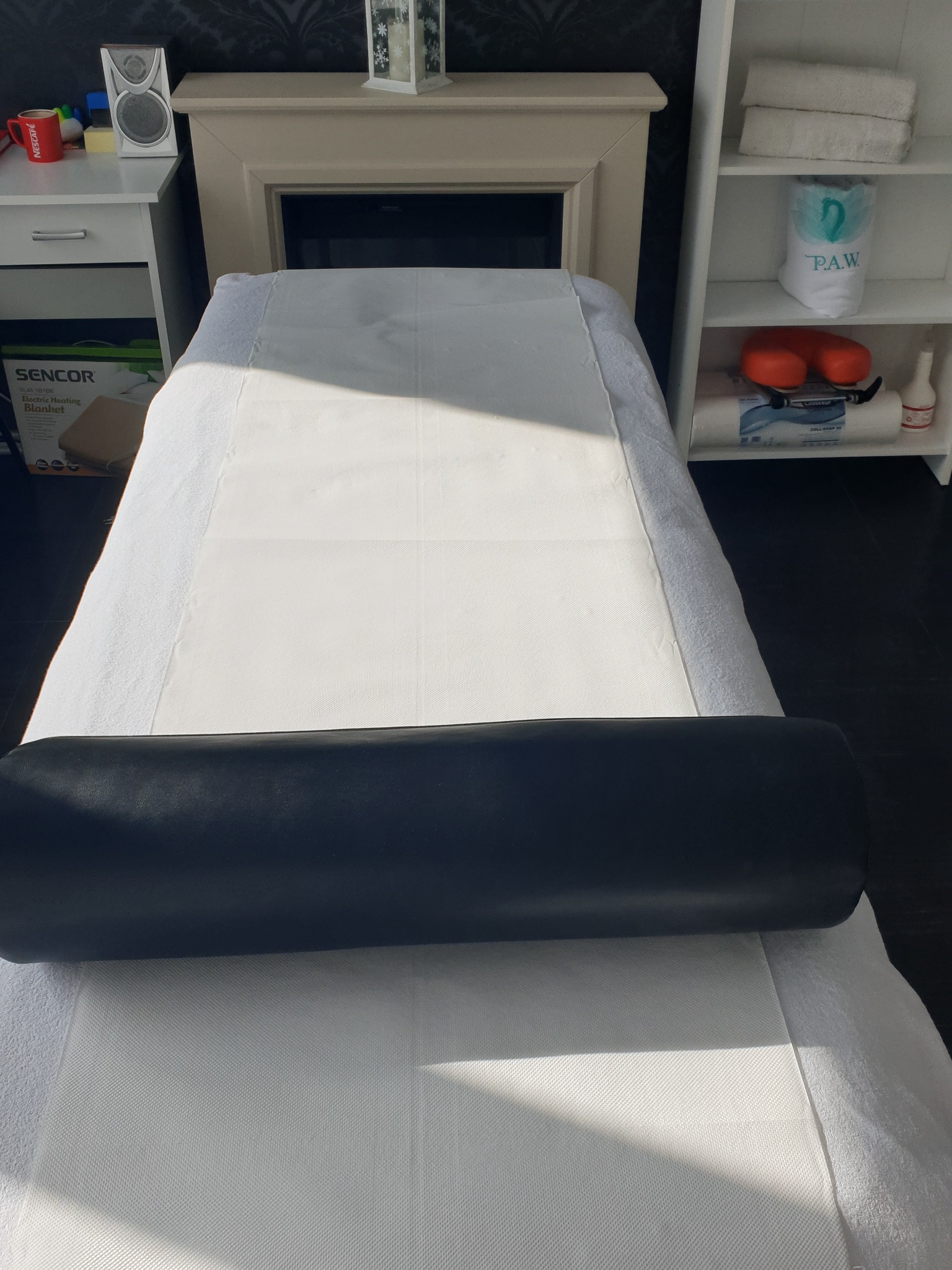 Przygotowane stanowisko do zabiegów: biały, profesjonalny stół z podkładem higienicznym, czarny wałek pod nogi, w tle regał z ręcznikami i kosmetykami.