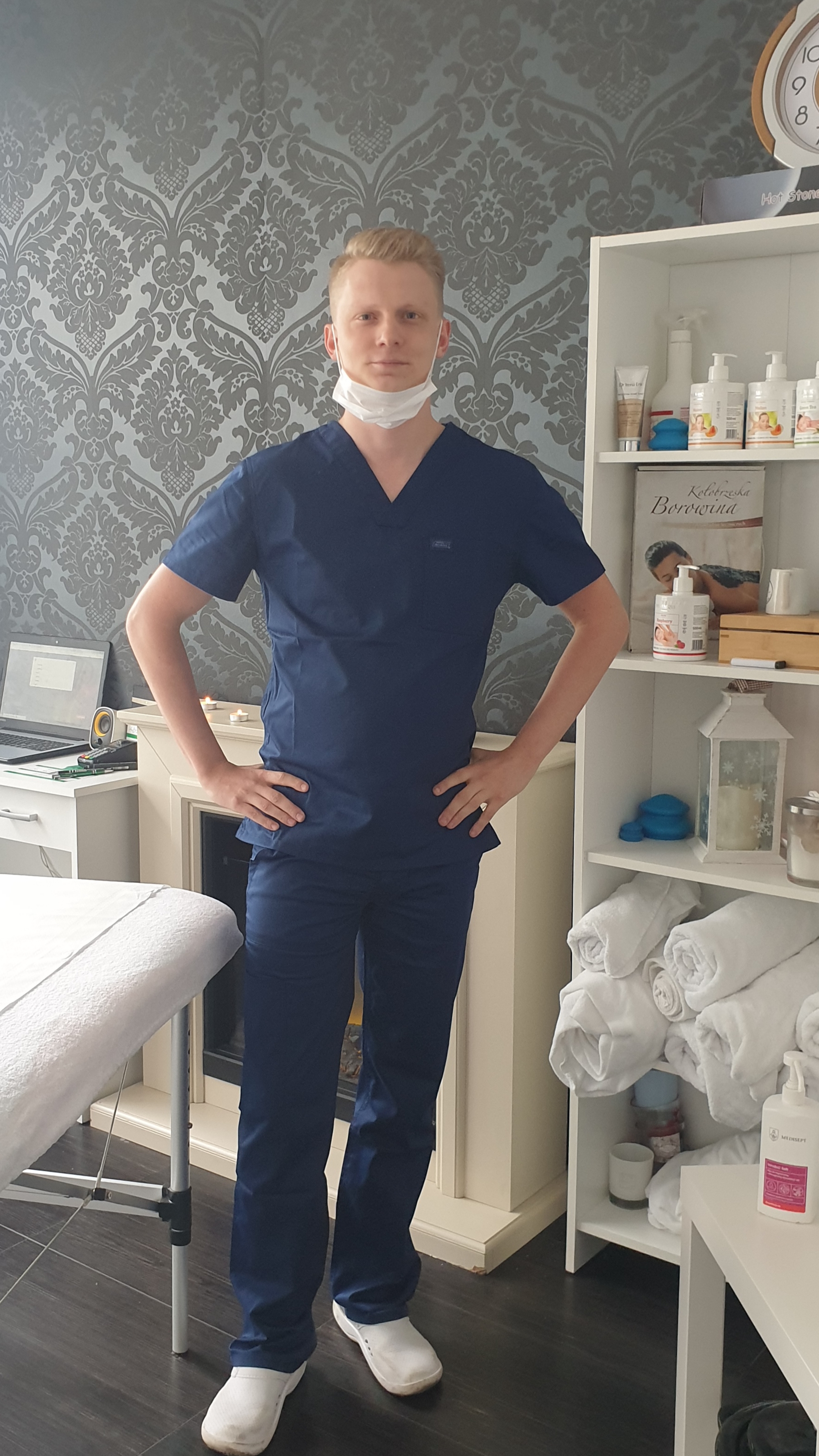 Młody mężczyzna w granatowym uniformie i maseczce ochronnej, stojący w gabinecie z tapetą w ornamenty, obok leżanki, kominka i regału z kosmetykami i ręcznikami.