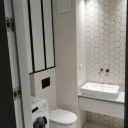 Nowoczesna, biała łazienka z heksagonalnymi płytkami na ścianie, umywalką nablatową i pralką widoczną przez uchylone drzwi.