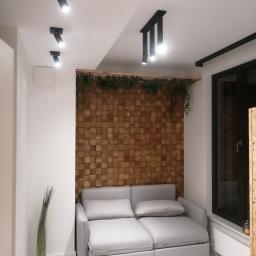 Przemek Łodożyński - Nowoczesny salon z drewnianą ścianą z kwadratowych paneli, oświetleniem punktowym na suficie i szarą sofą.