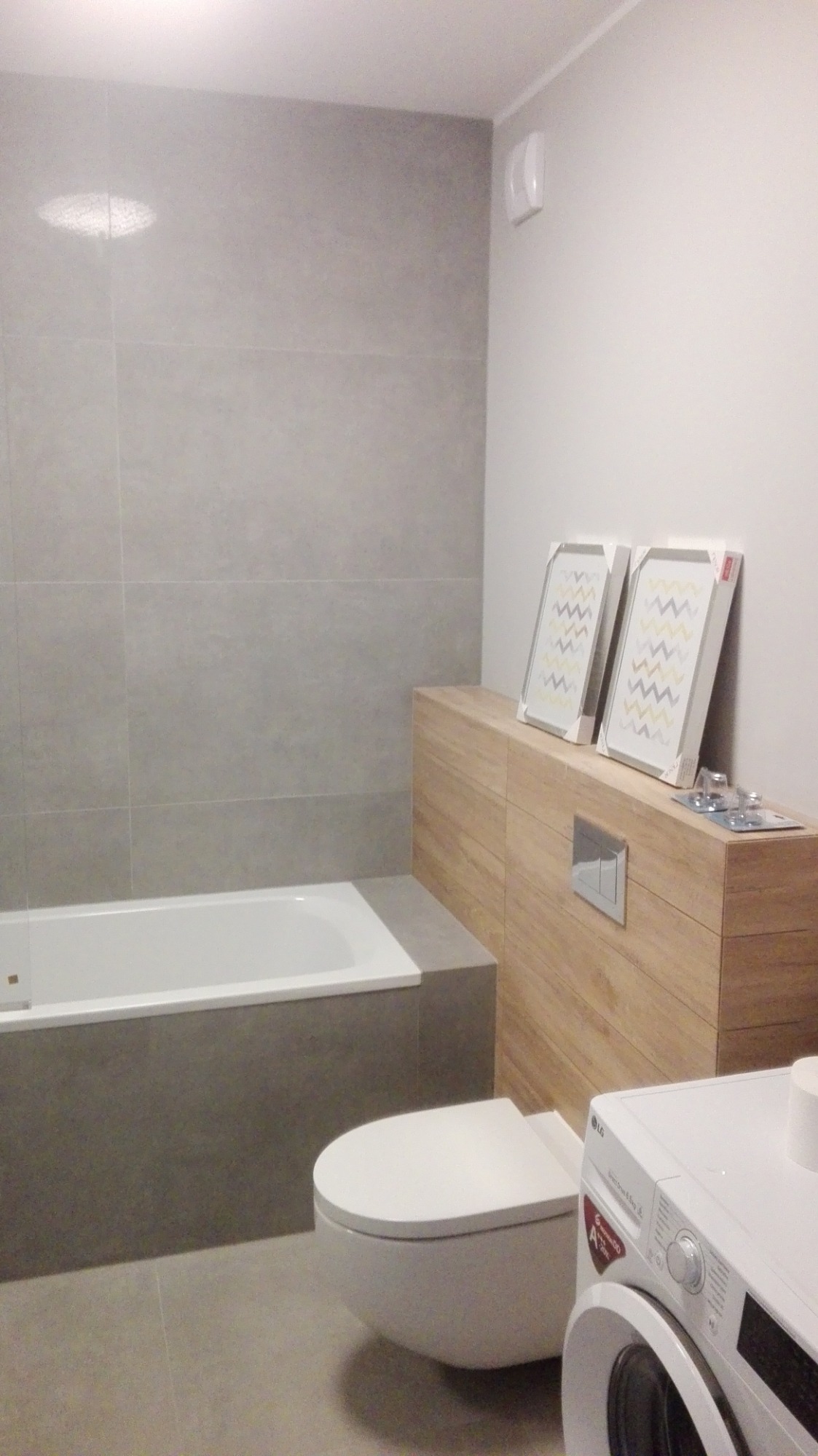 Nowoczesna łazienka z szarymi płytkami, białą wanną, podwieszaną toaletą, obudową z drewnopodobnych paneli, pralką i dekoracyjnymi obrazkami w ramkach na obudowie.