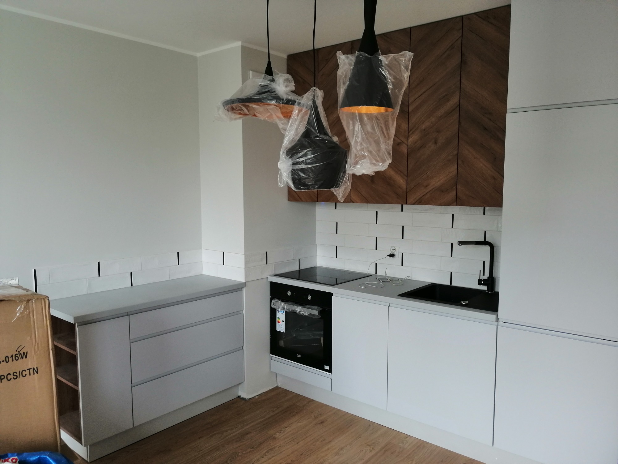 Nowoczesna kuchnia w trakcie montażu: białe szafki z drewnianą okładziną w jodełkę, czarny zlew i bateria, sprzęt AGD pod folią ochronną, wiszące lampy w osłonach.