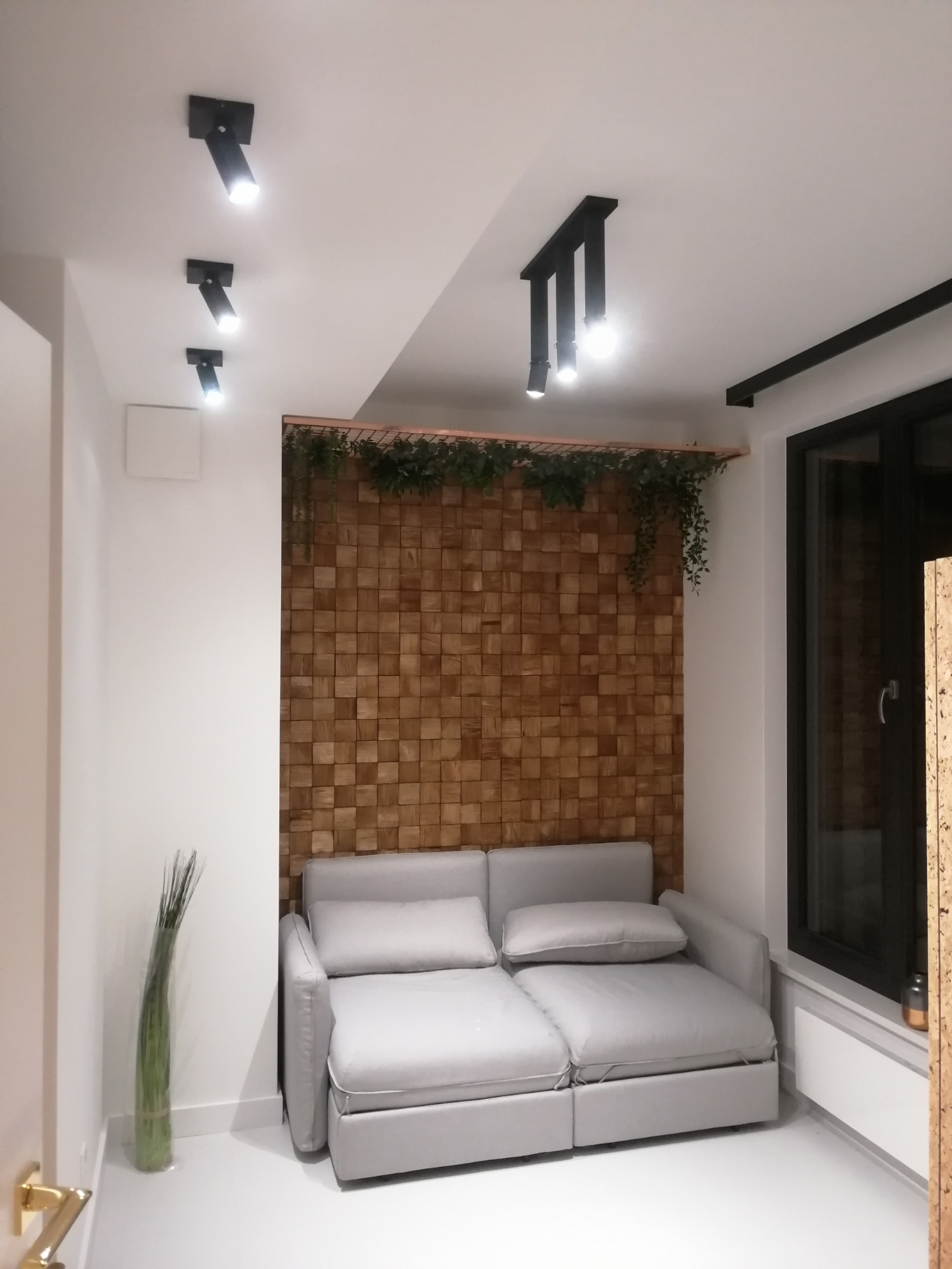 Nowoczesny salon z drewnianą ścianą z kwadratowych paneli, oświetleniem punktowym na suficie i szarą sofą.