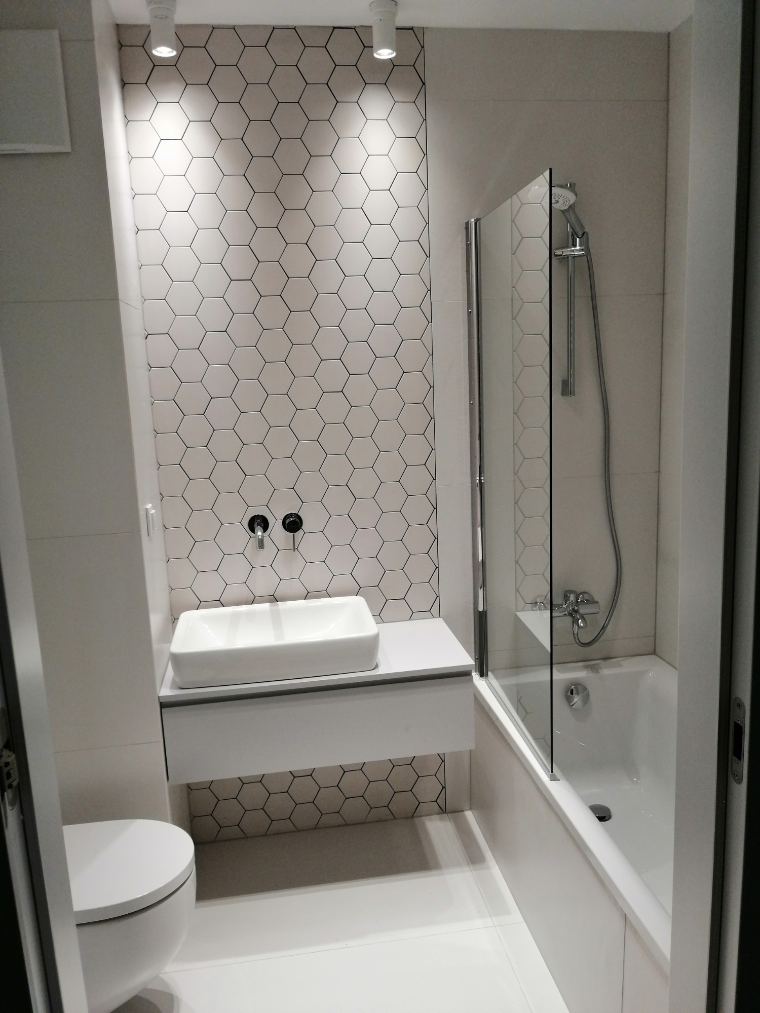 Nowoczesna łazienka w bieli i szarości z heksagonalnymi płytkami na ścianie, prostokątną umywalką nablatową, wanną z parawanem nawannowym i podświetleniem punktowym.