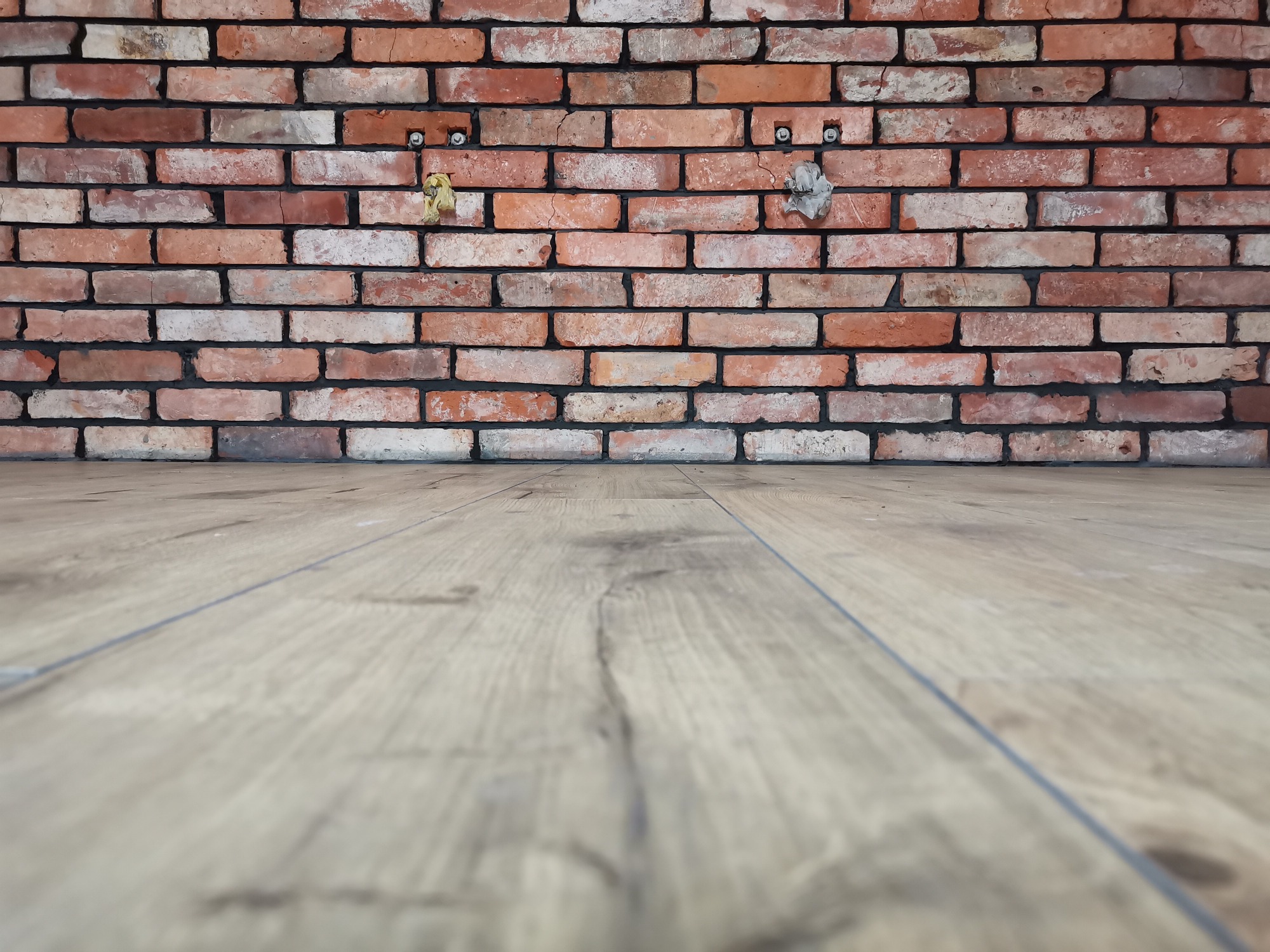 Czerwona ceglana ściana z widocznymi śladami po niedokończonych pracach, fragment drewnianej podłogi w dolnej części kadru.