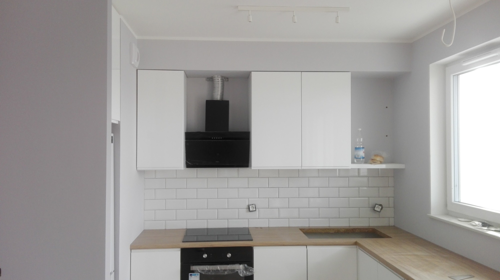 Nowoczesna kuchnia w szaro-białej tonacji z białymi szafkami wiszącymi, czarnym okapem, drewnianym blatem i białymi kafelkami na ścianie. Płyta indukcyjna i piekarnik jeszcze w folii ochronnej.
