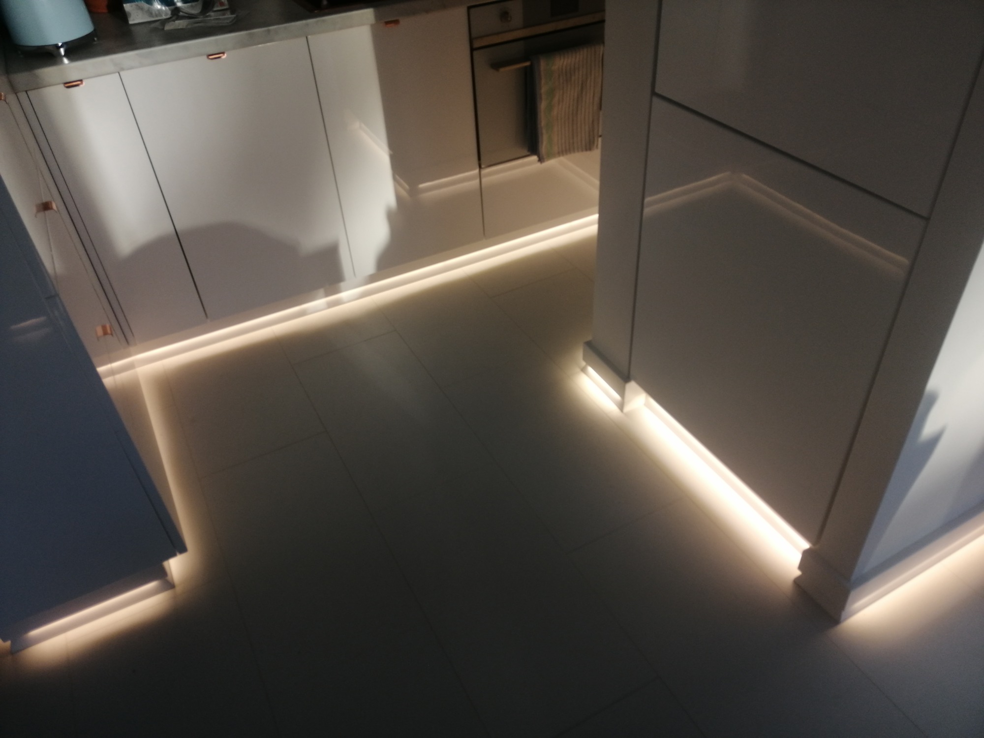 Nowoczesna kuchnia z białymi szafkami i podświetleniem LED pod szafkami dolnymi, oświetlającymi jasną podłogę.