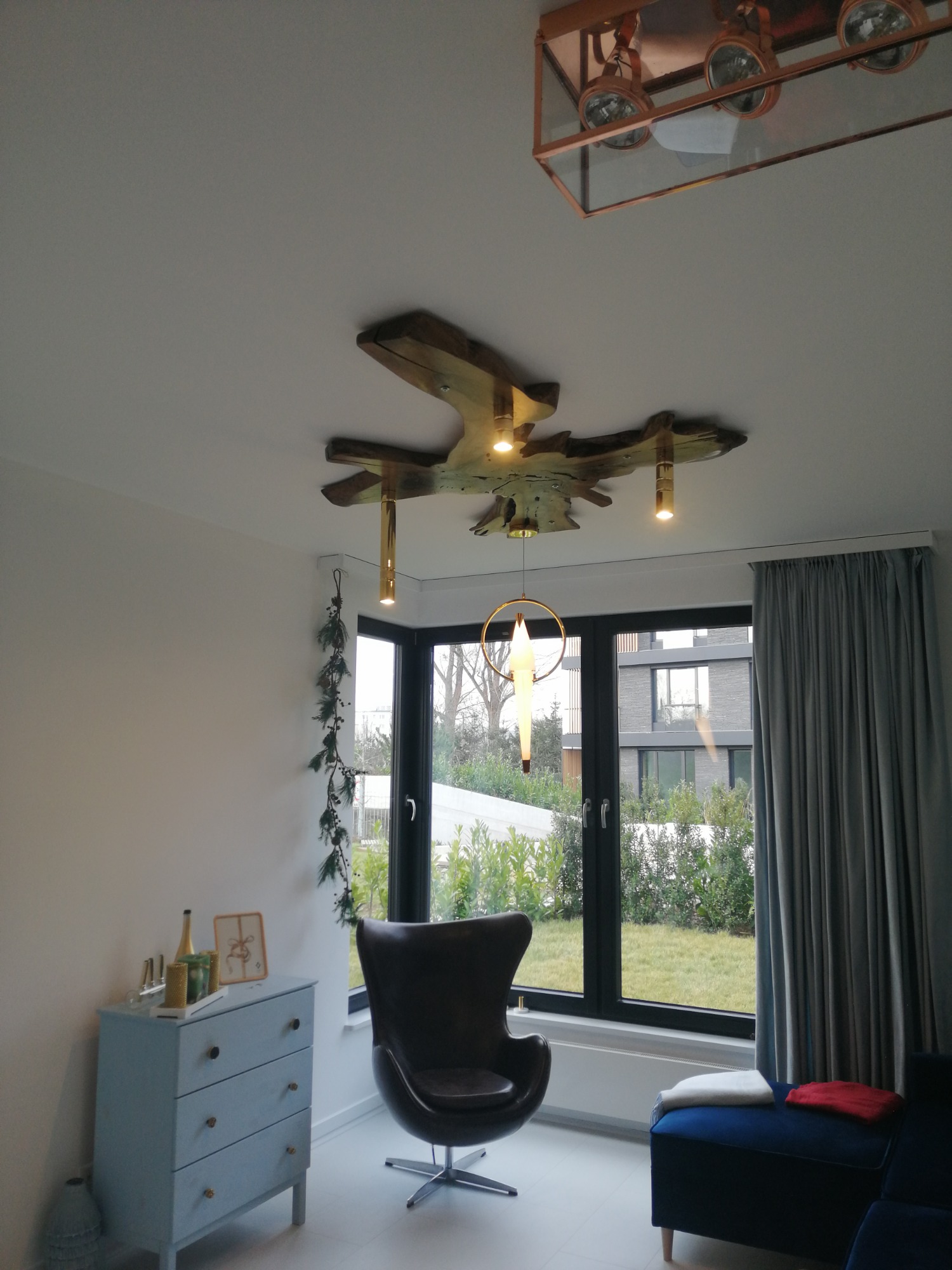 Nowoczesny salon z designerskim oświetleniem sufitowym: drewniana instalacja świetlna i lampa w geometrycznej oprawie, widok na zewnątrz przez duże okno.