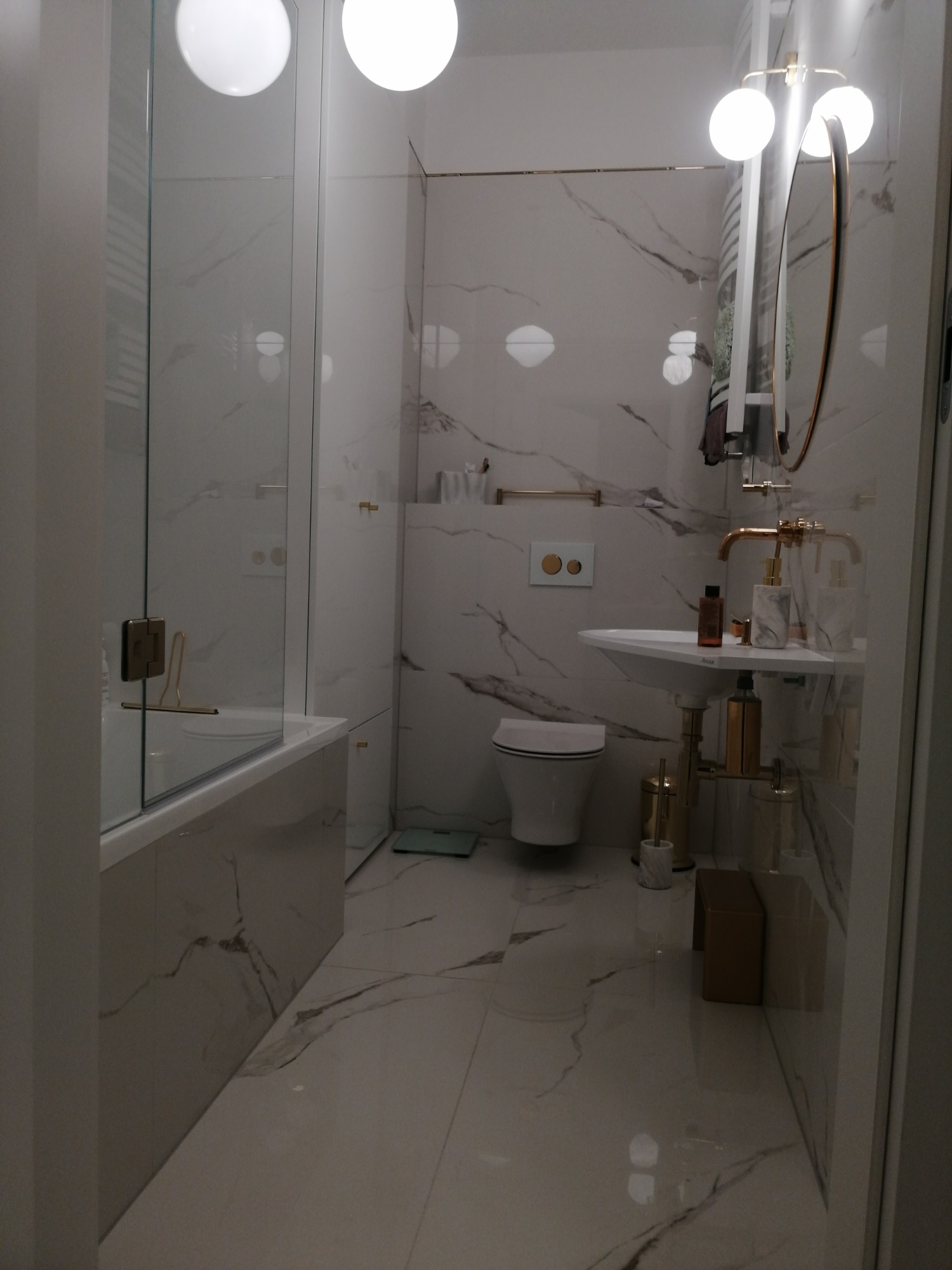 Nowoczesna łazienka z marmurowymi płytkami, złotymi akcentami i okrągłymi lampami sufitowymi, widok na kabinę prysznicową, umywalkę i toaletę.