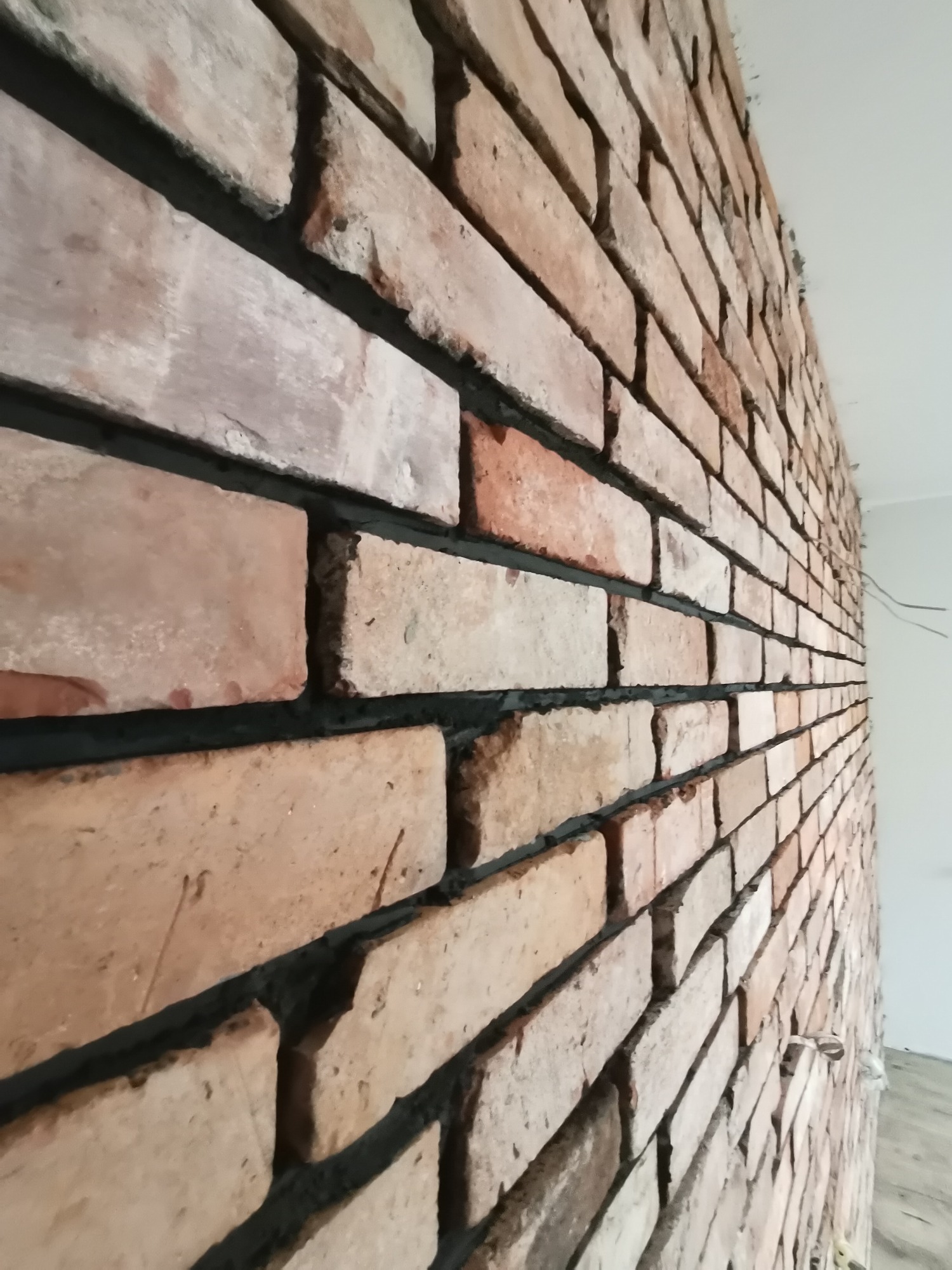 Ściana wyłożona cegłą klinkierową o zróżnicowanej kolorystyce, z widoczną czarną fugą, wykonana pod kątem, z fragmentem białego sufitu i wystającymi przewodami elektrycznymi.