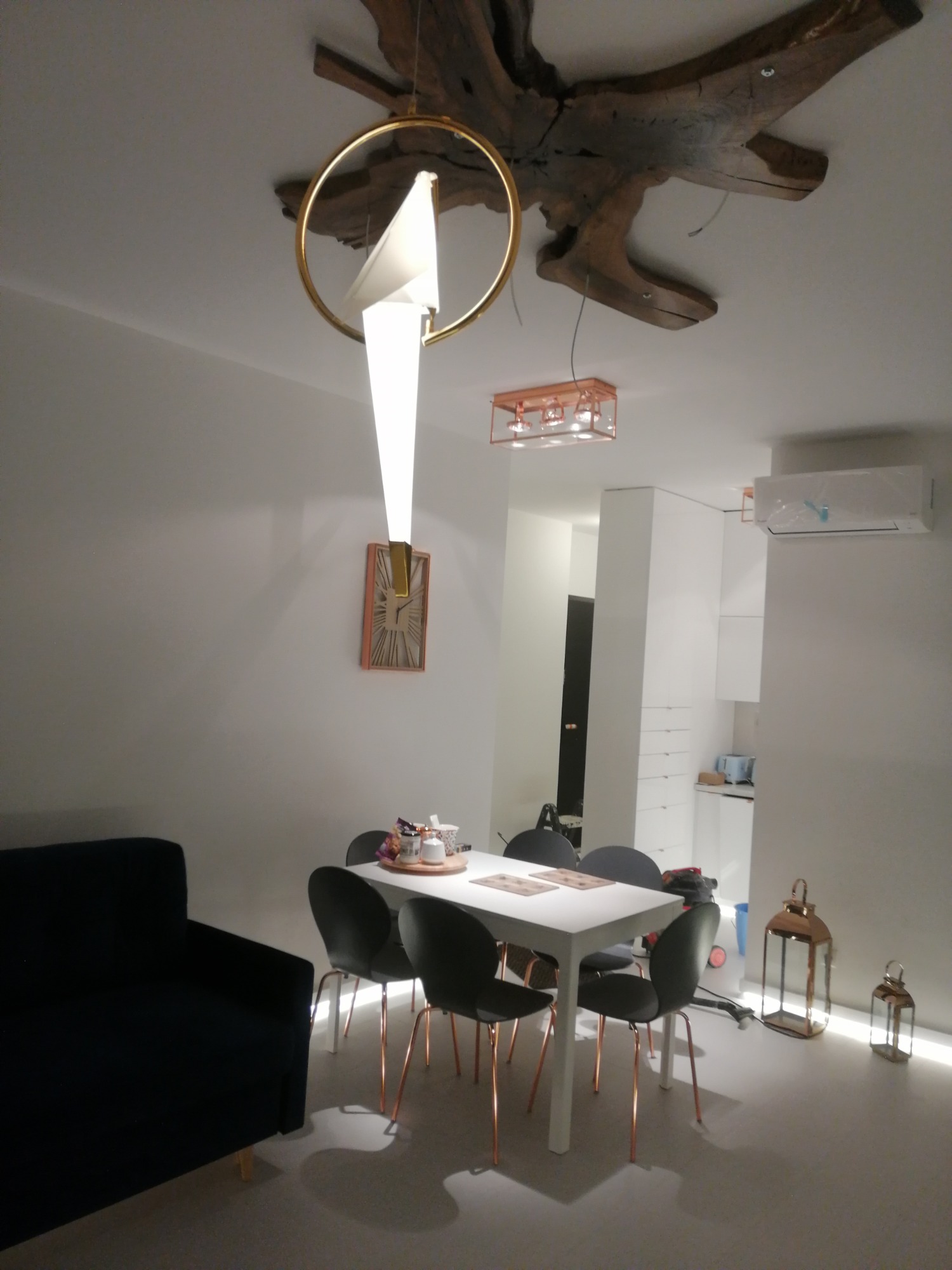 Nowoczesne wnętrze jadalni z czarną sofą, białym stołem otoczonym szarymi krzesłami na miedzianych nogach, designerską lampą z elementami drewna na suficie i dekoracyjnymi lampionami na podłodze.