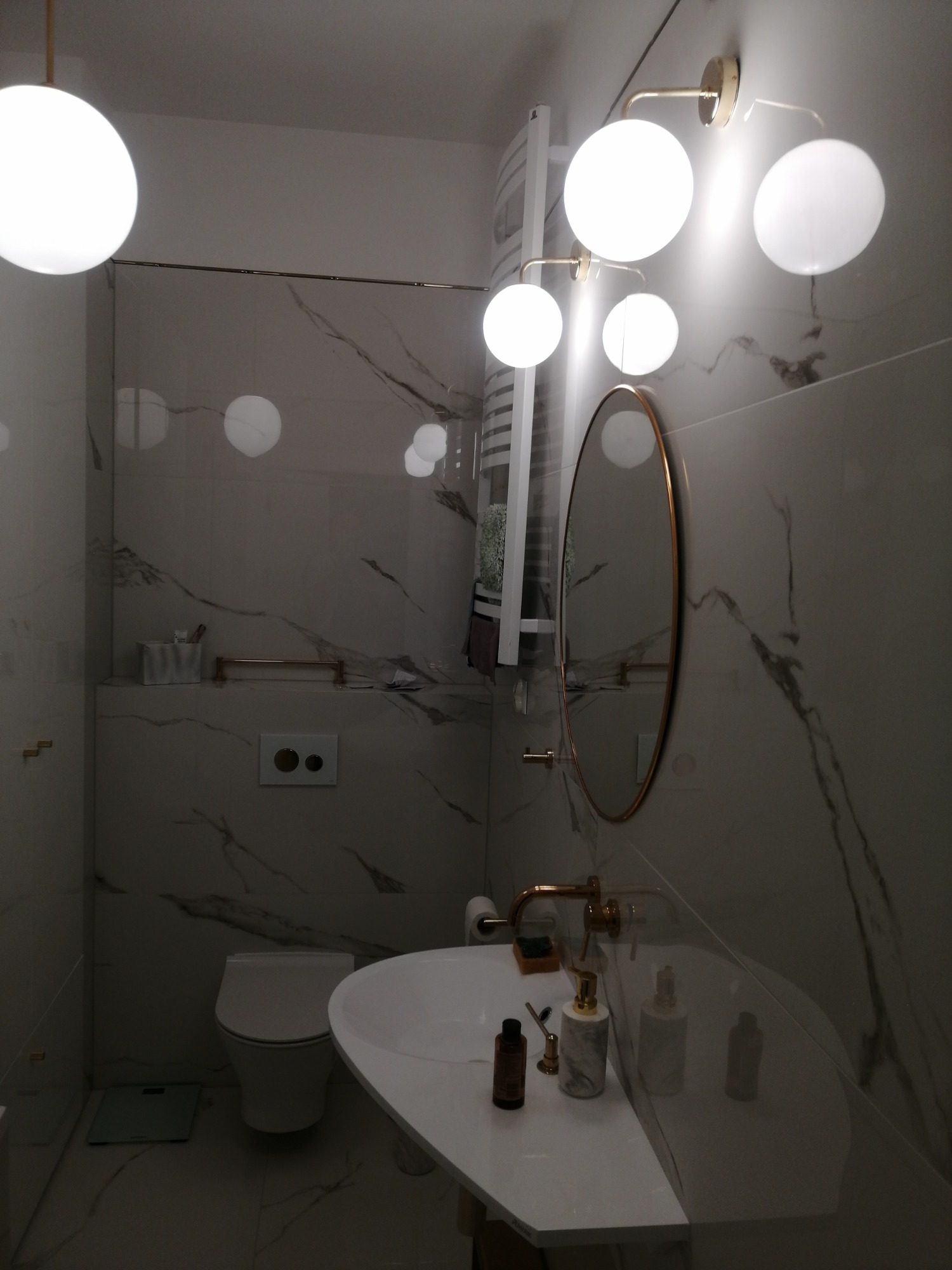 Nowoczesna łazienka w Banino z marmurowymi płytkami, owalnym lustrem w złotej ramie, kulistymi lampami i białą umywalką z dozownikami mydła.