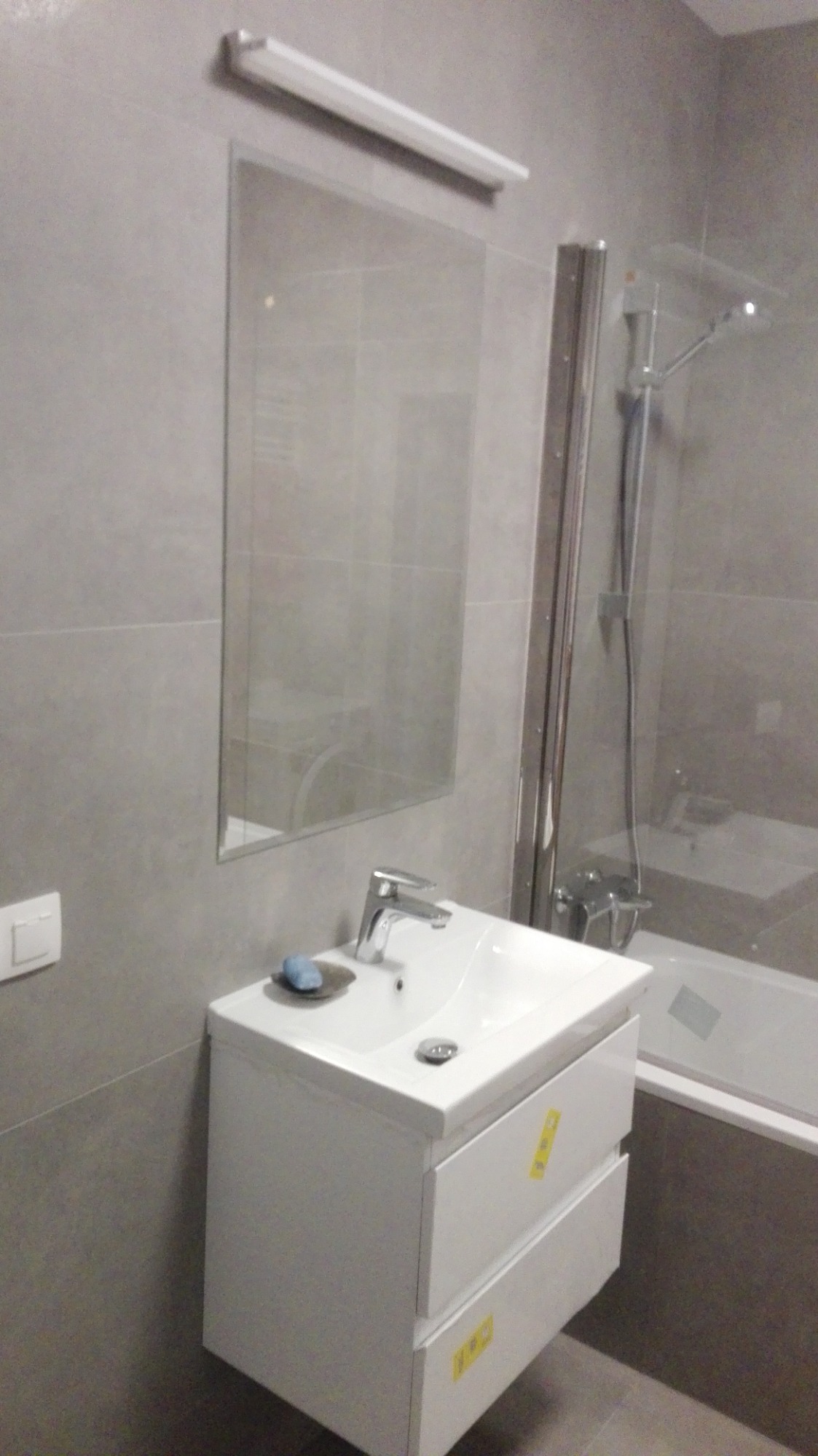 Szara łazienka z umywalką na szafce z dwiema szufladami, lustrem i kabiną prysznicową z widoczną baterią. Na umywalce leży mydło w mydelniczce.