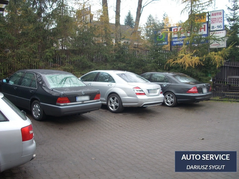 Trzy zaparkowane samochody Mercedes-Benz różnych generacji na tle szyldu warsztatu samochodowego z drzewami w tle.