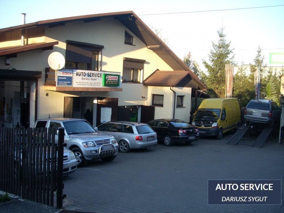 Budynek warsztatu samochodowego z zaparkowanymi autami, w tym jeden na podnośniku, oraz tablica reklamowa z logo Bosch i Castrol.