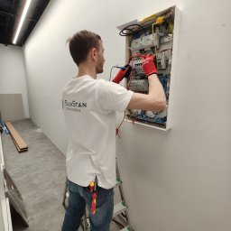 Sunstan Sławomir Stanisławek - Elektryk w czerwonych rękawicach, z miernikiem w dłoniach, sprawdza napięcie w skrzynce bezpiecznikowej w nowym budynku; na koszulce widoczne logo firmy SunStan.