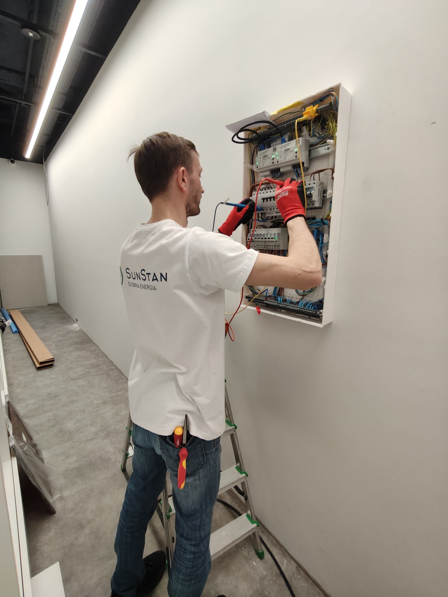 Elektryk w czerwonych rękawicach, z miernikiem w dłoniach, sprawdza napięcie w skrzynce bezpiecznikowej w nowym budynku; na koszulce widoczne logo firmy SunStan.