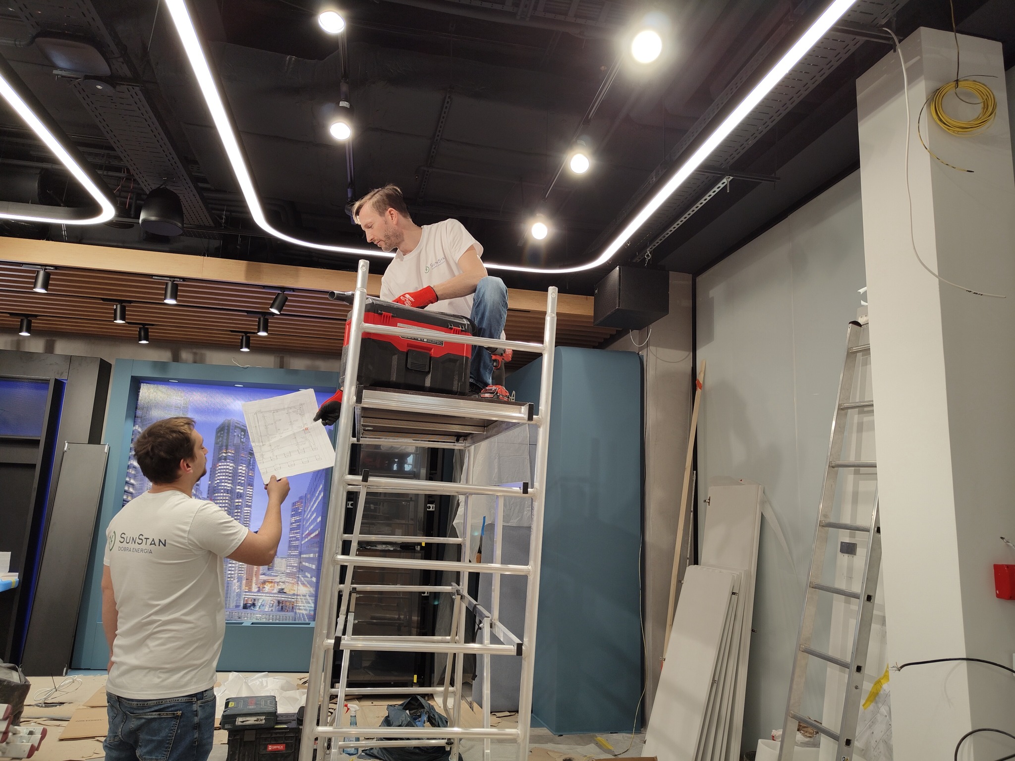 Dwóch mężczyzn podczas montażu oświetlenia w lokalu w Raszynie. Jeden na aluminiowym rusztowaniu z czerwonym pojemnikiem na narzędzia, drugi trzyma plan instalacji. Widoczne przewody i elementy...