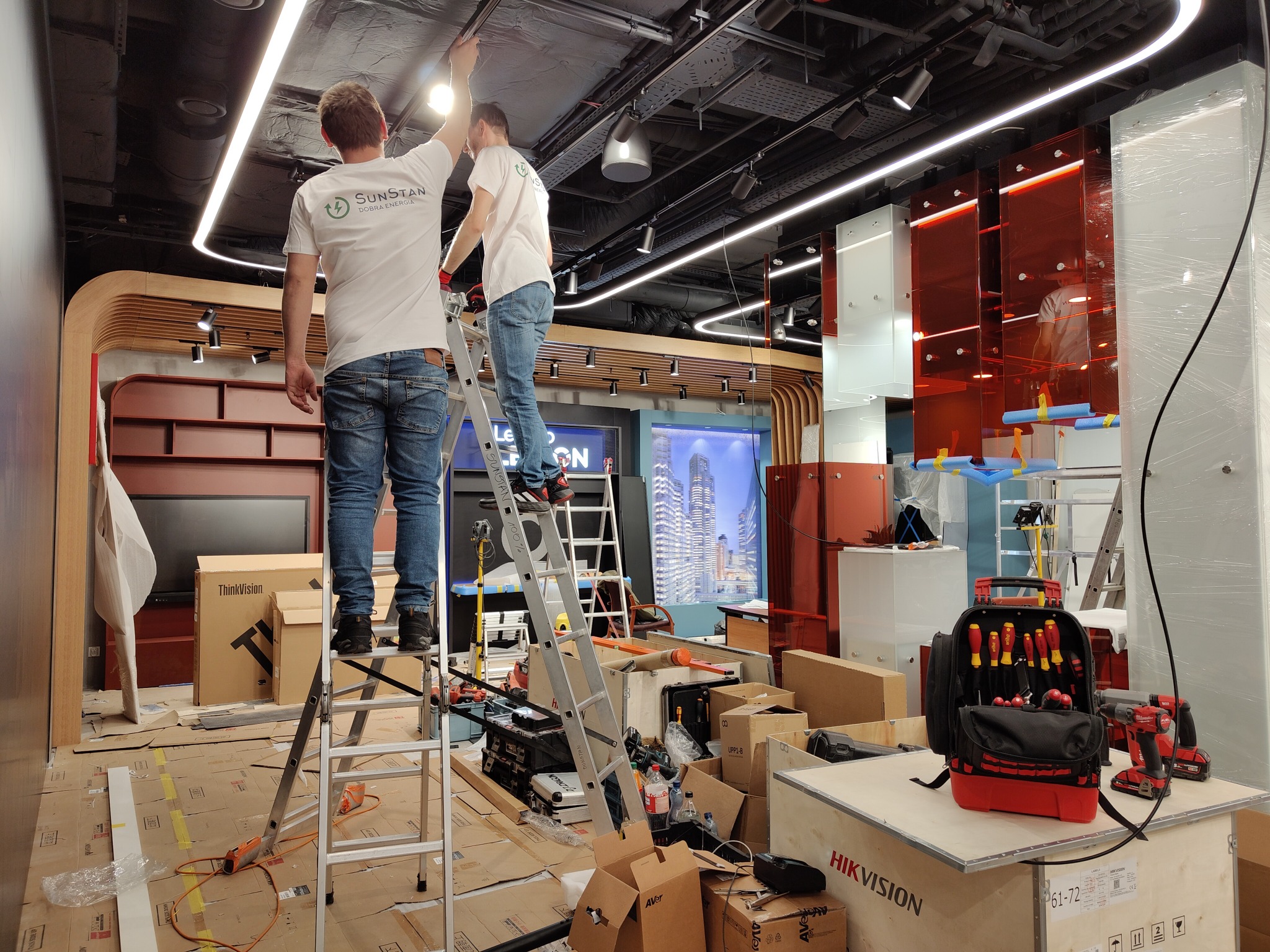 Dwóch monterów na drabinach instaluje oświetlenie LED na suficie w nowoczesnym biurze, widoczne narzędzia i kartony z elektroniką, logo firmy na koszulkach.