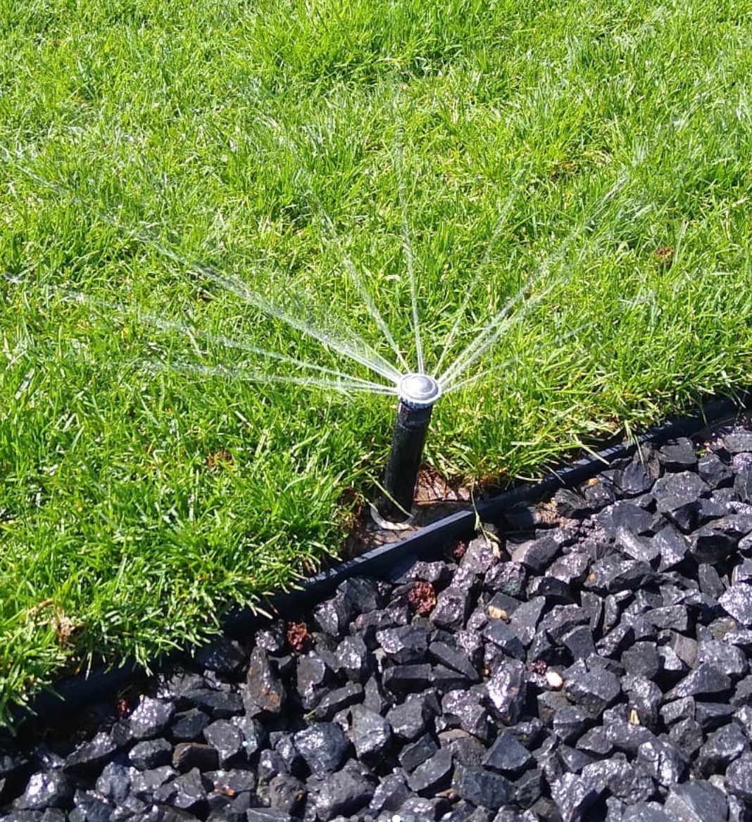 Automatyczny system nawadniania trawnika z dyszą rozpryskującą wodę, otoczony czarnym kamieniem dekoracyjnym i zieloną trawą.