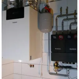 Biały kondensacyjny kocioł gazowy marki Buderus z szarym zbiornikiem wyrównawczym i instalacją hydrauliczną na tle białych płytek, widoczne rury wentylacyjne i logo firmy w rogu.