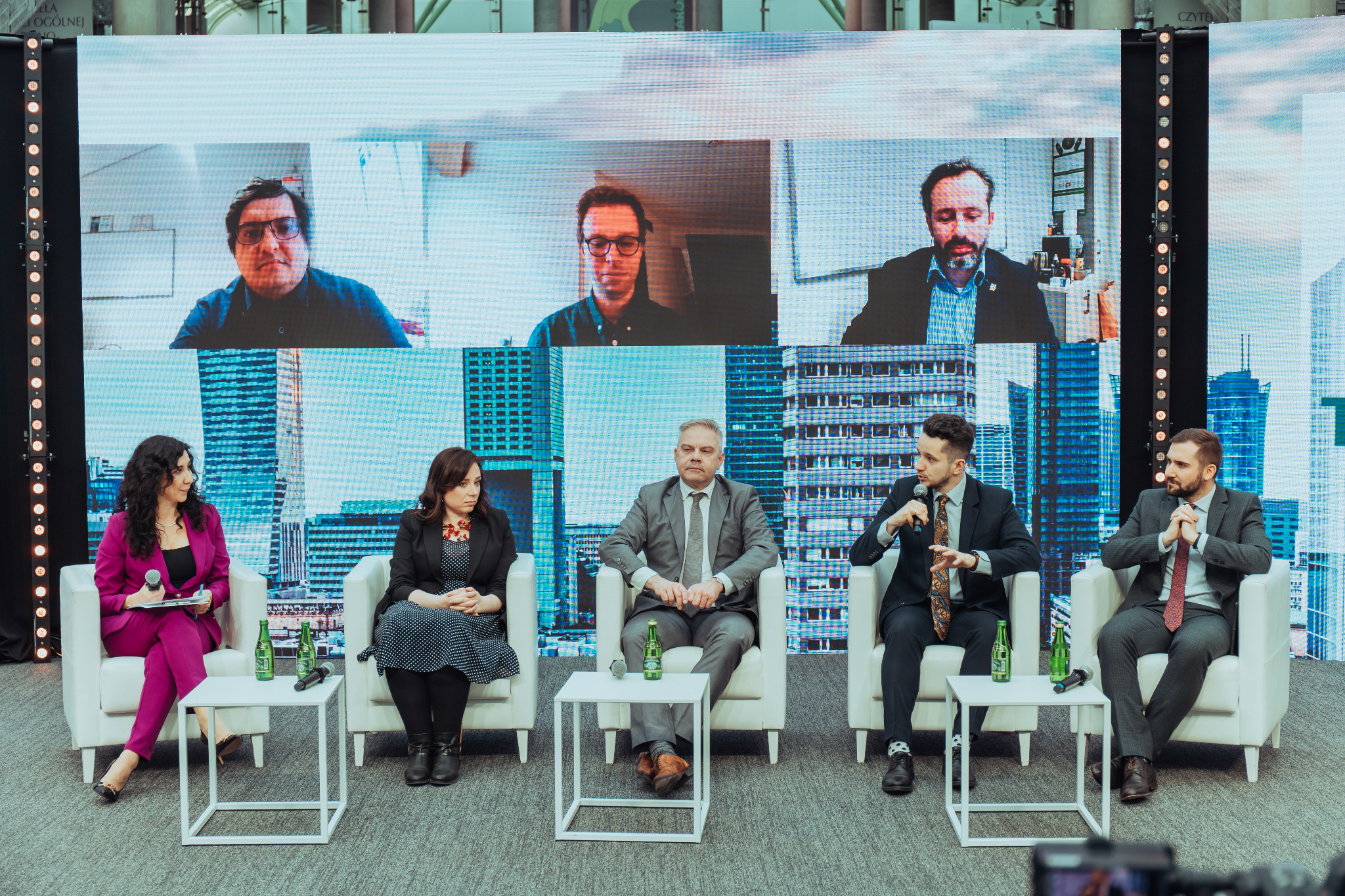 Panel dyskusyjny podczas konferencji z uczestnikami na żywo i zdalnymi, siedzący na białych krzesłach na tle ekranu LED z wirtualnym tłem miejskim.